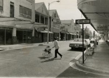 A woman with a child in a stroler crosses a street with storefronts on either side and a one-way sign.