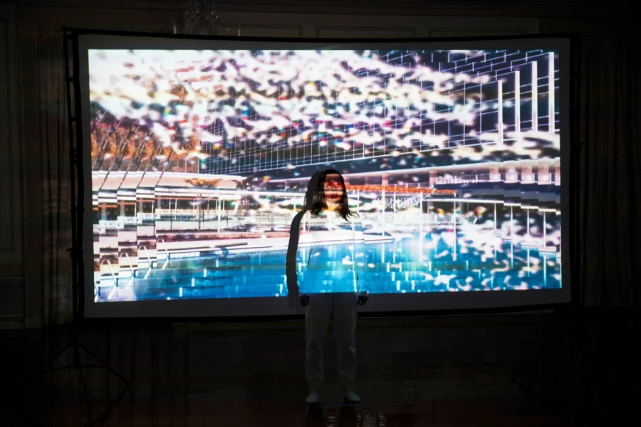 An Asian woman dressed in white stands in front projected images from Venezuelan from archival sources with visual effects 