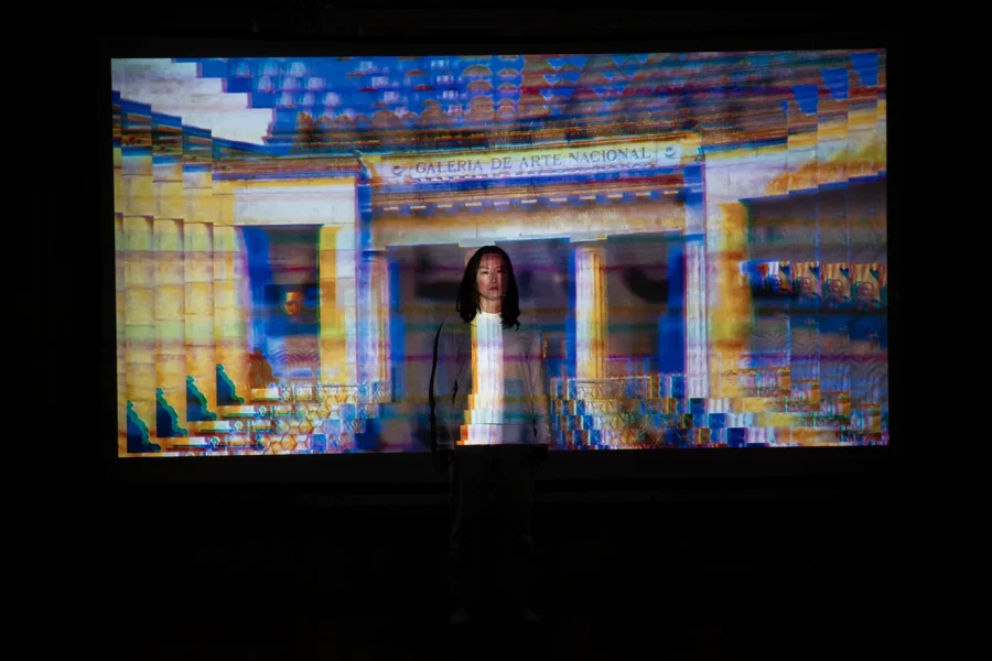 An Asian woman dressed in white stands in front projected images from Venezuelan from archival sources with visual effects 