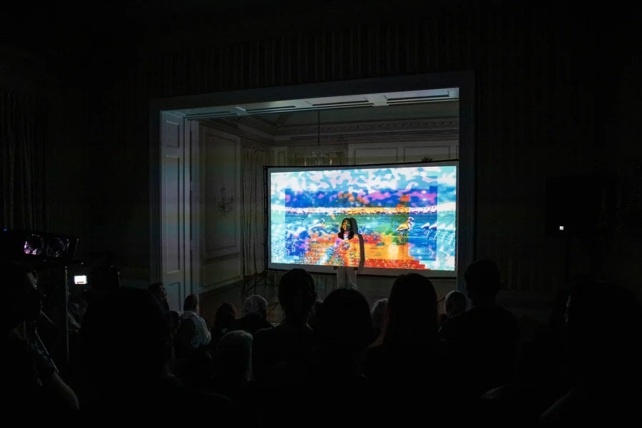 An Asian woman dressed in white stands in front projected images from Venezuelan from archival sources with visual effects 