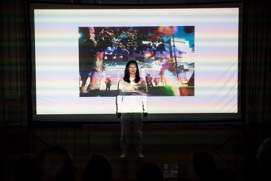 An Asian woman dressed in white stands in front projected images from Venezuelan from archival sources with visual effects 