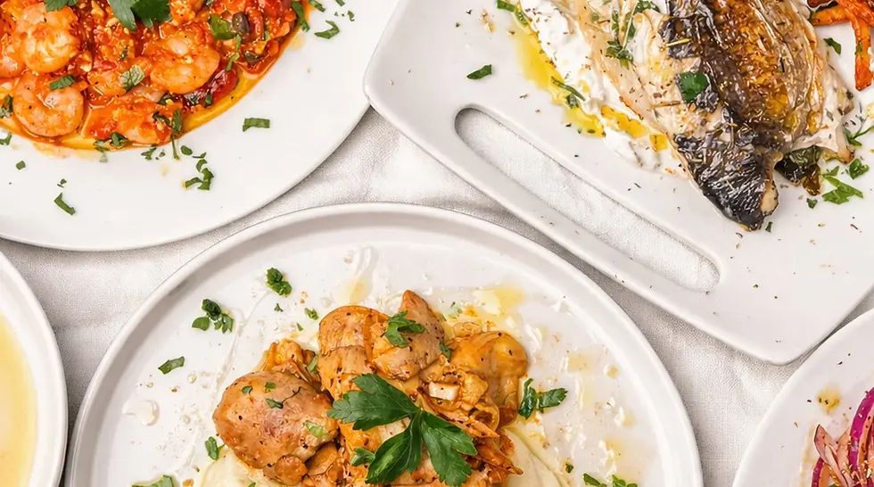 Overhead view of plated seafood dishes—shrimp, fish, and herb garnish—on white plates, bright restaurant-style spread.