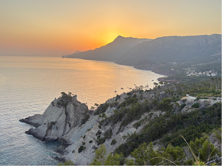 Sunrise over a rocky coastal bay with cliffs, calm water, and distant mountains under a golden orange sky.