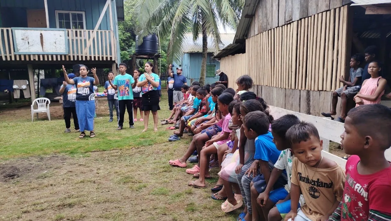 Children seated beside a wooden building during an outdoor group activity, in Greg’s campaign photograph.