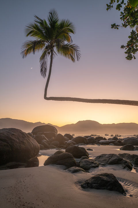 Fotografia de praia de Angra dos Reis com o nascer do sol dourado e a lua decrescente, com pedras em primeiro plano na orla