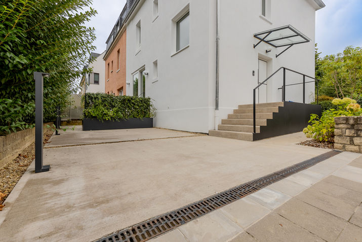 Entrée bâtiment moderne, marches béton, allée pavée, verdure.