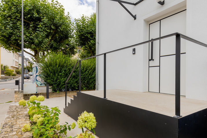 Entrée moderne, escaliers et garde-corps noirs, façade blanche.
