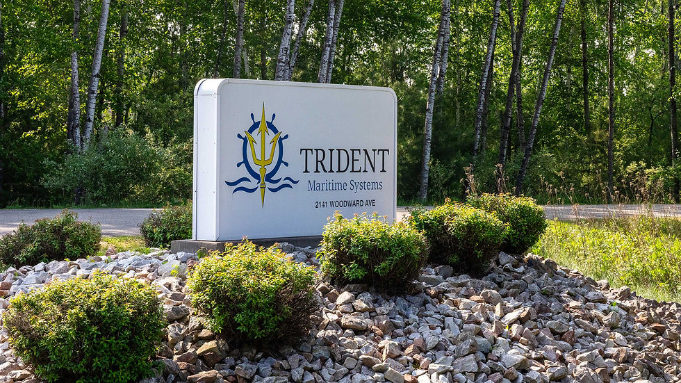 Sign for Trident Maritime Systems with logo in a wooded area, surrounded by rocks and greenery. Text: "2141 Woodward Ave."