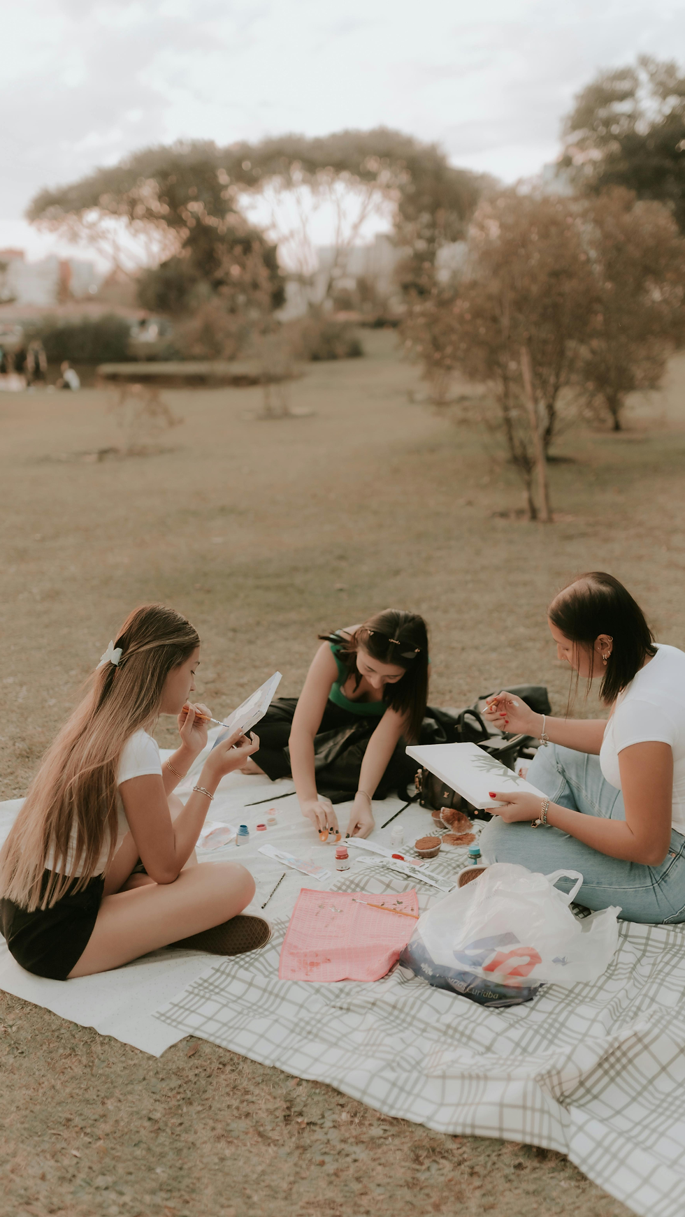 Three women are painting on canvases outdoors on a plaid blanket in a park. Art supplies are scattered. The mood is relaxed.