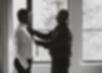 Two men in a room; one adjusts the other's tie. They're near a large window with trees outside. The mood is focused and collaborative.