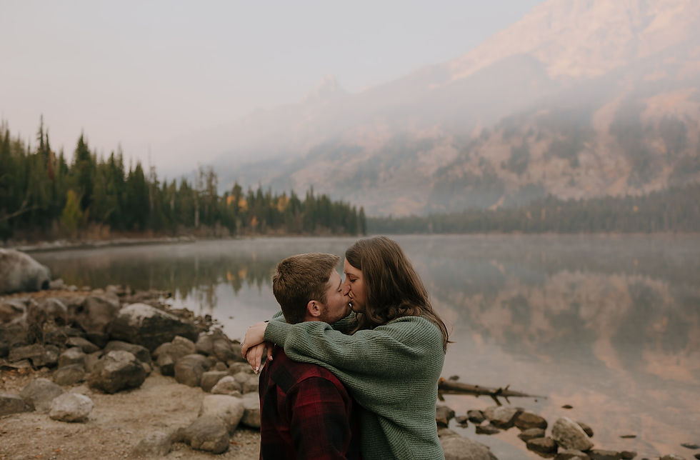 A couple kisses by a serene lake with mountains and trees in the background. The mood is romantic and peaceful, with earthy tones.