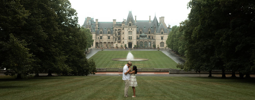Couple embracing on a lawn in front of a grand mansion with a fountain, surrounded by trees. Overcast sky, creating a romantic mood.