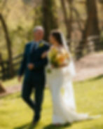 A bride in a white dress and veil holds a bouquet, walking with an older man in a blue suit, smiling in a sunny, wooded outdoor setting.