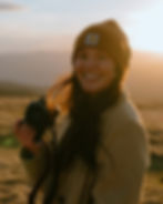 Woman smiling with a camera at sunset, wearing a brown beanie with logo. Fields and golden light in the background, creating a warm mood.