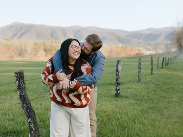 A couple embraces joyfully in a grassy field with mountain views. The woman in a wavy-patterned sweater and glasses laughs warmly.
