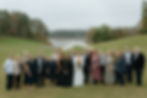 A large group in formal attire poses on a grassy hill by a lake. A bride holds flowers, smiling. Trees and water are in the background.