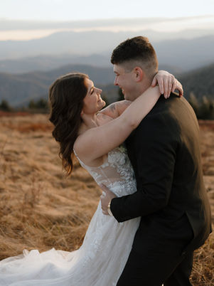 Couple embracing joyfully in a field at sunset. Woman in white dress, man in black suit. Mountains and soft clouds in the background.