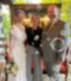 Bride in lace dress, groom in gray suit, and photographer smile amid floating bubbles. Greenery and neon sign in background. Joyful mood.