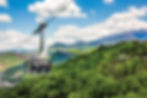 Cable car gliding over lush green forest with mountains in the background under a blue sky with fluffy clouds, creating a serene view.