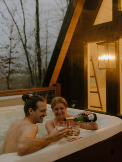 a couple shares a bottle of champagne in front of an a-frame cabin, while soaking in a hot tub