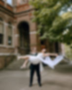 A groom in a suit lifts a bride in a white dress and veil outside a brick building. Both wear sunglasses, exuding joy beneath leafy trees.