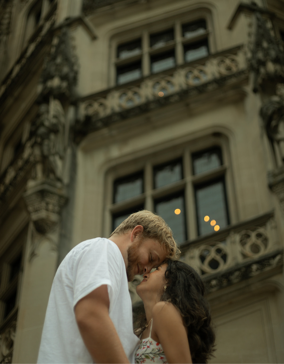 A couple embraces, gazing into each other's eyes, in front of an ornate building. She wears a floral dress; he wears a white shirt. Romantic mood.