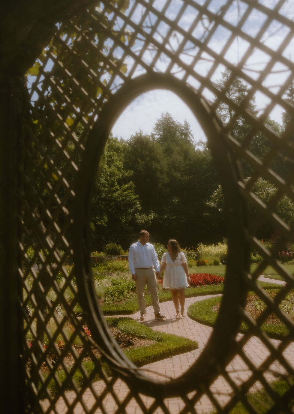 A couple holds hands, walking in a garden viewed through a lattice window. They're surrounded by lush greenery and colorful flowers.