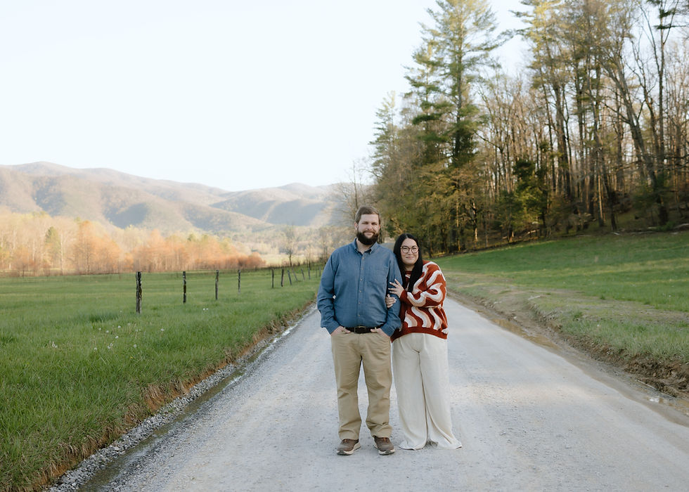 Couple stands on a rural dirt road with mountains and trees in the background. The man wears a blue shirt; the woman, a red patterned sweater.