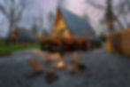Cozy A-frame cabin with lit string lights and fire pit. Wooden chairs surround the fire on a gravel patio. Warm, inviting evening scene.