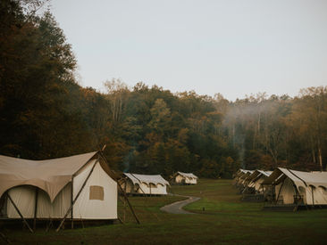 Rows of canvas tents in a grassy campsite, surrounded by autumn trees. A winding path leads through the misty, serene setting.
