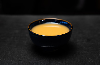 Bowl of soup, dark blue with golden-brown liquid, sitting on dark surface.