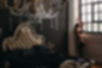 Woman in black lingerie poses by a large window in a luxurious bedroom with dark walls, ornate bed, and crystal chandelier.