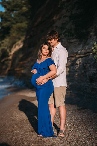 pregnant woman wearing blue dress stands on a serene beach embraced by a man with a white shirt as they hold hands over her swollen bump