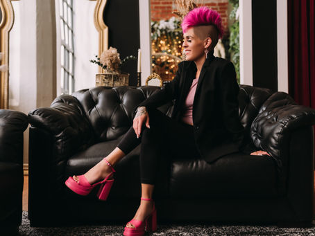 Person with bright pink mohawk sits on black leather sofa in stylish room. Wears black suit, pink shoes. Decor includes gold mirrors, plants.