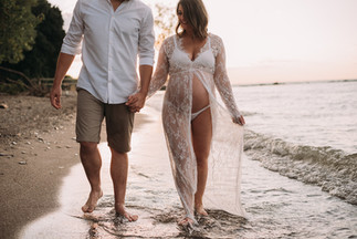 Man in white shirt and pregnant woman in lace dress hold hands while walking down a shoreline at sunset, conveying intimacy and serenity, with waves in the background.