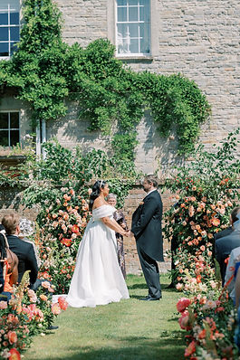 Outdoor wedding ceremony at Elmore Court's walled garden with bride and groom standing in front of a broken floral arch and an aisle lined with floral meadows of coral peonies and roses.
