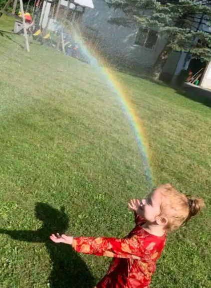 Photo of a small child outside holding out her hand towards a rainbow in the spray from a garden hose.