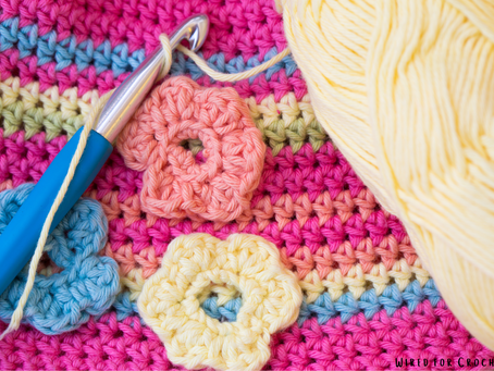 A multicoloured crochet blanket with a hook, 3 crochet flowers and a yellow yarn ball placed on top.