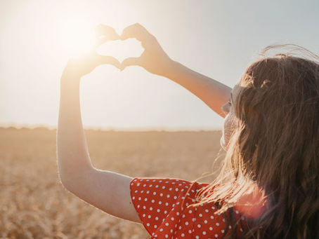 A woman standing in a golden field at sunset forms a heart shape with her hands around the sun, symbolizing optimism, warmth, and connection in Leading with Sunshine Yellow Energy.