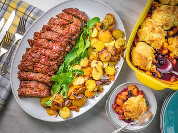 Sliced meatloaf with arugula and roasted potatoes on a white plate. Cobbler with fruits in yellow dish. Plaid napkin and cutlery nearby.