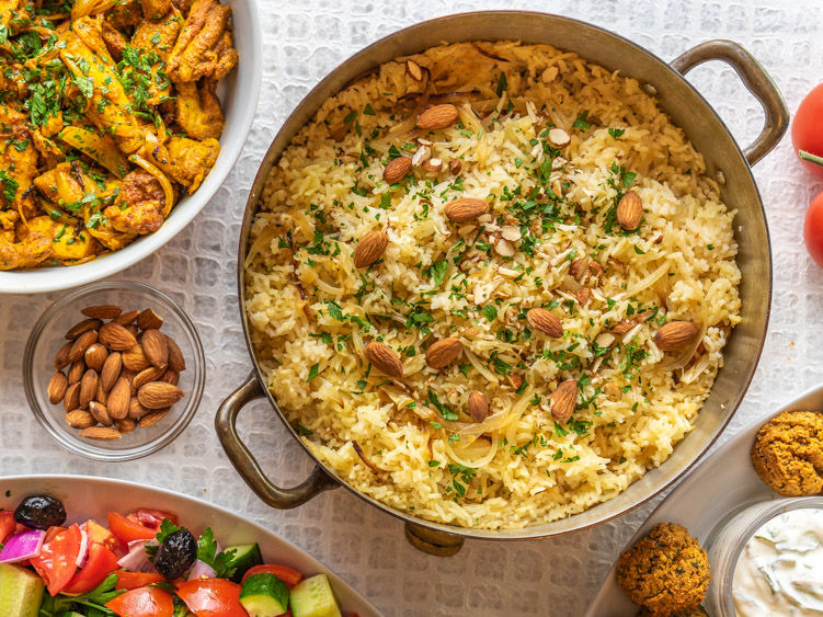 A bowl of rice with almonds and herbs, alongside spicy chicken, almonds, salad, and falafel. Background features a textured tablecloth.