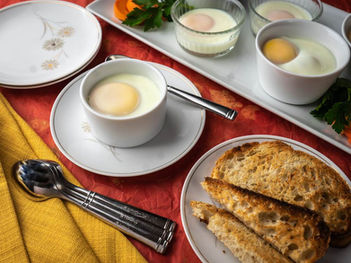 Eggs in white cups and toasted bread on plates, set on a red and yellow tablecloth. Spoons and parsley garnish add detail.