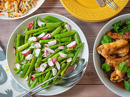 Bowls of vibrant asparagus salad with radish, golden chicken wings, and colorful noodles on a wooden table. Yellow plate and utensils nearby.