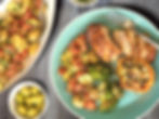 A plate with roasted chicken, potatoes, tomatoes, broccoli, and stuffed pepper. Green olives in a bowl. Wooden table background. Bright colors.
