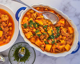 A casserole dish with vibrant stew of chickpeas, meat, and squash, topped with greens. A spoon is in the dish. Another dish and bowl nearby.