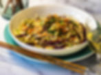 Colorful noodle salad with veggies in a white bowl on a marble table. Wooden chopsticks and a glass of white wine are nearby.