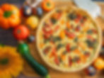 Quiche with tomatoes and pesto on a wooden board, surrounded by colorful tomatoes, zucchini, bell pepper, garlic, and a sunflower.
