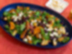 Colorful roasted vegetable salad with arugula and feta on a blue platter, set on a red tablecloth. A side dish of shaved cheese nearby.