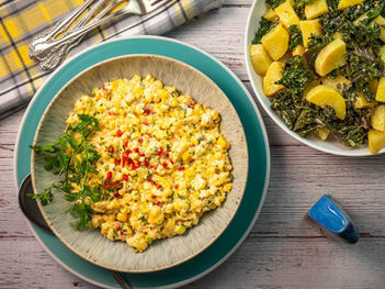 Scrambled eggs with red peppers and herbs on a plate, beside potatoes with kale. Silverware on a plaid cloth. Rustic wood table.