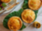 Golden pies on green leaves, served on a wooden table with a fork, knife, and salad. A small jar of mustard is beside them. Cozy vibe.